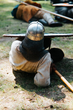 A soldier wearing a chainmail and helmet lies on the ground in a grassy area during a medieval festival