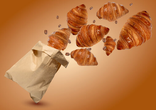 Fresh golden croissants and coffee beans fly out of a craft paper bag on a warm brown background.