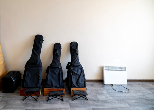 Musical instrument cases standing against a plain interior wall next to a portable electric heater and a black amplifier placed on a wooden floor in a minimalist room with natural light illumination