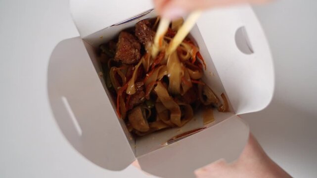 Woman eating Chinese udon noodles with meat and vegetables from a takeout box using chopsticks on a white background