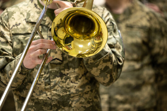 Armed forces personnel in modern camouflage uniforms play a brass trombone instrument detailed patches and rank insignia while performing during a public event near an urban municipal building and
