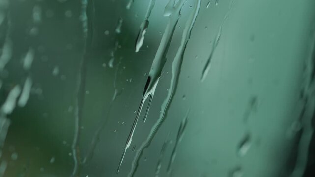 Large water drops and rain streaks on a greenish-blue glass surface. Close-up macro shot with a blurred background and atmospheric natural light.