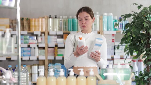 Young woman holds moisturizing creams with collagen in hands, she reads the composition and chooses between in two options against the background of the shelves with medicines in the pharmacy. High
