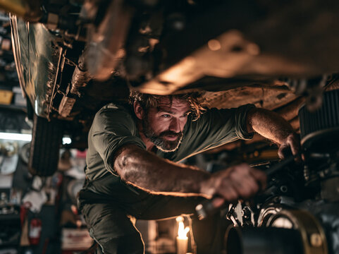 A focused mechanic with a beard is working underneath a car in a garage, his hands holding a wrench AI generative