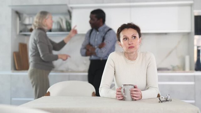Upset middle-aged woman sitting in the kitchen and man and mature woman arguing standing behind 