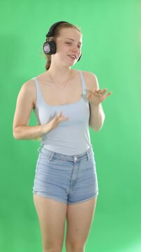 Young woman in casual clothes listens to music in modern headphones with high-quality sound on a chroma key background. Girl who is engaged in dancing enjoys her favorite song on a green background