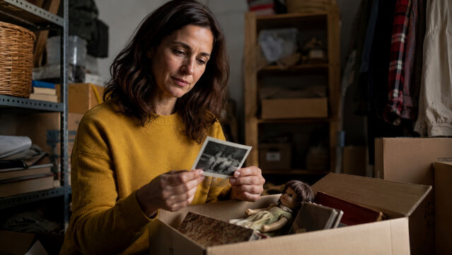 Emotional woman reflecting on past memories, holding treasured black and white photograph discovered in cardboard box of mementos and antique doll, sorting through personal history in dim storage room