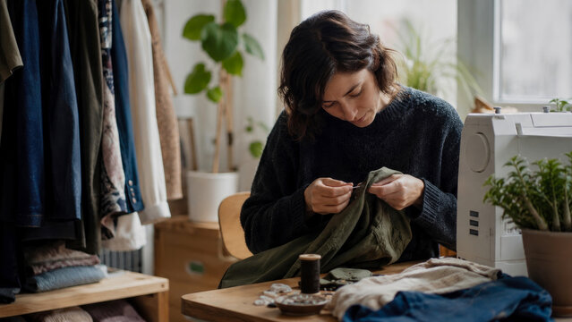 Diligent woman expertly hand-sewing and mending garment at her inviting home studio desk, surrounded by textiles and sewing machine, promoting sustainable slow fashion and creativity.
