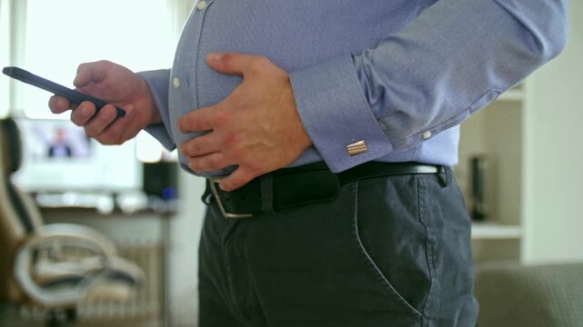 Fat man in shirt stroking his belly and use smartphone.