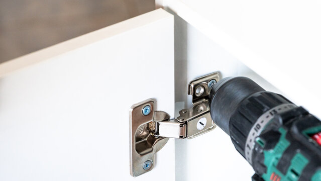 Close-up of a worker installing and adjusting cabinet door hinges using a power drill and screwdriver. Furniture assembly, maintenance, and DIY home repair concept.