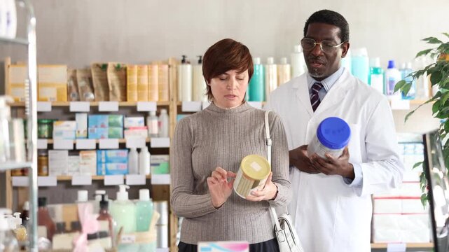 Woman choose buy infant food at pharmacy, compare with other good. Client check for absence of harmful substances, view product in details. African man pharmacist offer available assortment item