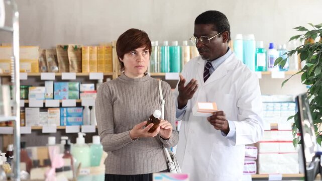 Woman choose buy antitussive syrup at pharmacy, compare with other good. Client check for absence of harmful substances, view product in details. African man pharmacist offer available assortment item