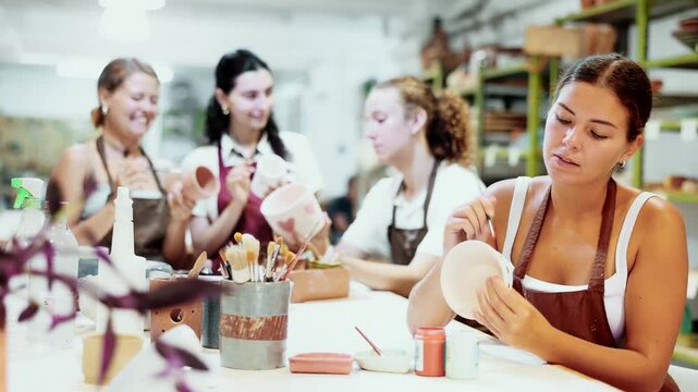 Young woman sits near table and decorates clay ceramic craft vase, plate, works in company of like-minded people. Hand made art, hobbies and creativity. 