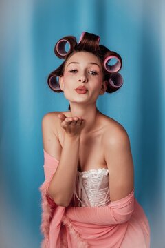 Blowing a kiss while wearing hair rollers and a pink robe in a studio setting
