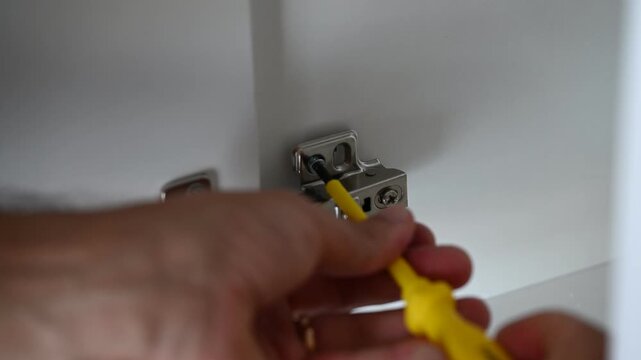 Close-up video of a technician installing and adjusting cabinet door hinges using a screwdriver and power drill. Furniture assembly, repair, and maintenance concept