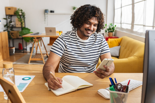 Young adult Indian man student studying with smartphone and notebook at home desk. Focused learner combining mobile technology and online education resources. Education and e-learning concept.