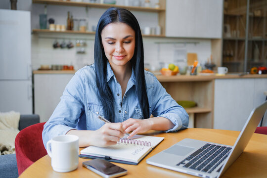 Woman working on laptop at desk while writing ideas and checklist in notebook, planning, creativity, organization and comfortable home office lifestyle concept