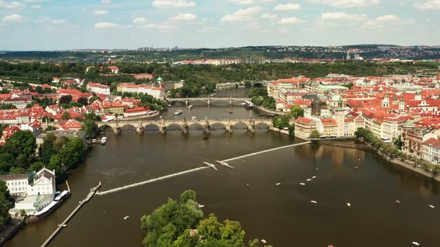 Charles Bridge spans Vltava river across historic Prague cityscape. Classic red tiled roofs fill Old Town district skyline. Small boats cruise past urban riverfront on bright summer day.