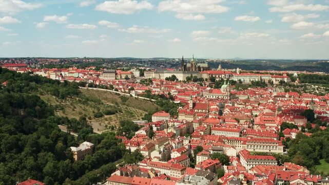 Prague Castle complex and Saint Vitus Cathedral rise above historic red tile roofs in Mala Strana district. European capital city panorama under sunny blue sky creates vibrant travel background.