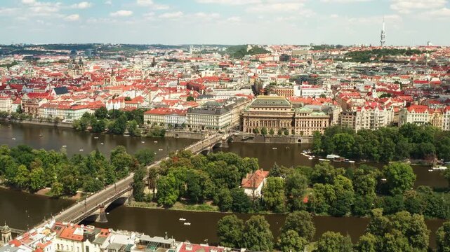 Prague cityscape featuring Vltava river, National Theater and historic bridge. Red tile roofs fill urban panorama under sunny blue sky. European capital city travel destination background.