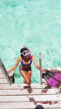 Woman after snorkel swim in paradise coral reefs ocean coming out of water removing fins to relax on overwater balcony. Raw unedited vertical video of real unretouched woman model without any makeup