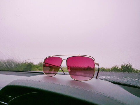 Pink tinted sunglasses rest on car dashboard. Raindrops visible on windshield behind. Car interior with red trim details. Outdoors view through wet glass. Reflections show trees and sky