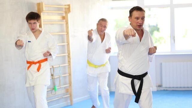 Motivated sporty man wearing white kimono and black belt performing kata routines to hone martial arts skills in training room with group of karate practitioners