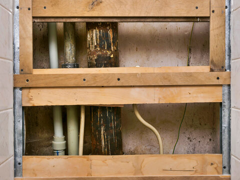 Exposed plumbing and wooden framing in tiled bathroom wall. Rusty pipes and exposed fixtures show renovation progress. Brown water filter attached to wall below frame