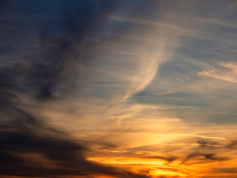 Dramatic sky at sunset with streaky clouds. Warm orange and yellow hues glow near horizon. Darker clouds contrast with soft light. Sky texture shows natural movement