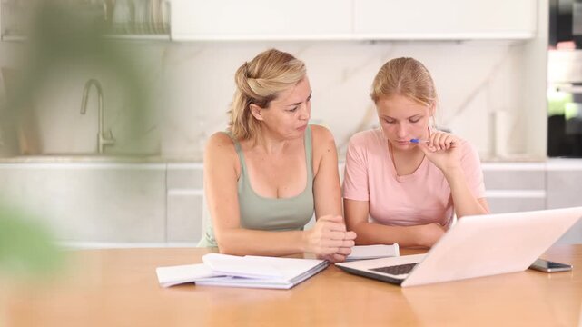 Family doing homework together. Mother assisting her teenage daughter sitting at table at home with school assignment. Diligent girl looking at laptop and writing in notebook