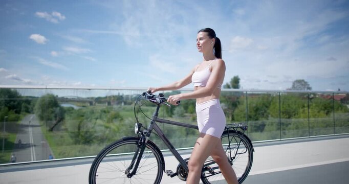 Young beautiful athletic woman in sportswear walking and riding a bicycle in the city in an urban park with a bridge and benches