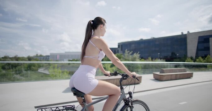 Young beautiful athletic woman in sportswear walking and riding a bicycle in the city in an urban park with a bridge and benches