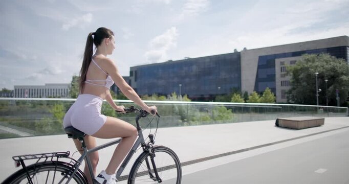 Young beautiful athletic woman in sportswear walking and riding a bicycle in the city in an urban park with a bridge and benches