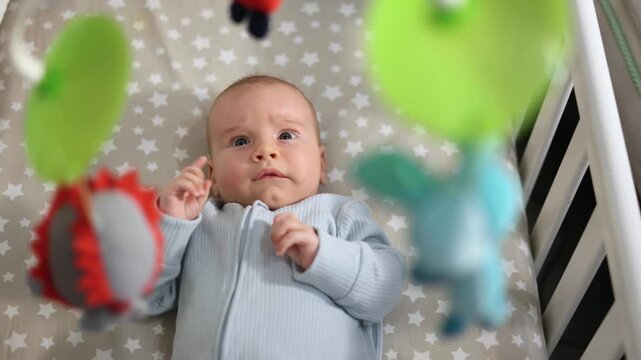 Baby lying in crib and looking up at hanging colorful mobile toys in a cozy nursery. 4k footage.