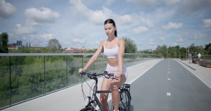 Young beautiful athletic woman in sportswear walking and riding a bicycle in the city in an urban park with a bridge and benches