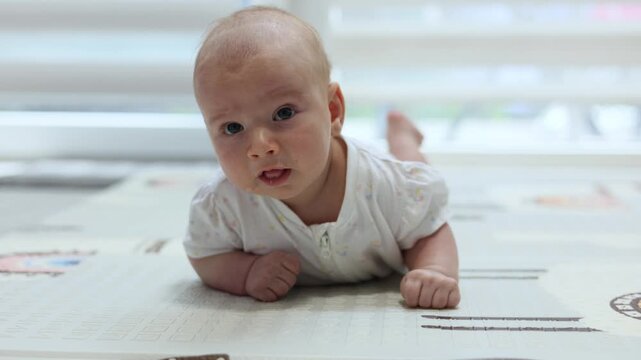 Adorable infant lying on tummy on a rug, looking curiously at the camera during tummy time at home. 4k footage.