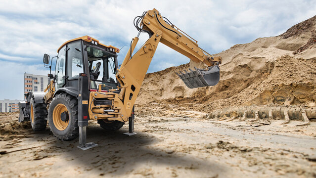 Universal excavator loader works on dirt at construction site with mounds of earth and buildings visible in background during cloudy day