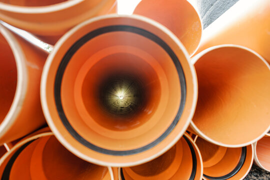 Workers are seen at a construction site with orange pipes stacked together. The sunlight shines through the opening of the pipes creating interesting patterns