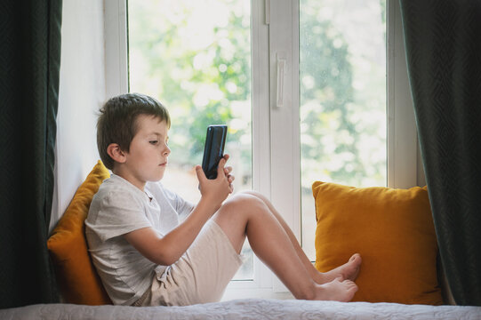 A 6-7 year old child sits on a windowsill at home with a mobile phone and plays. Mental health issues, unlimited gadgets, FOMO, harm to the child's psyche.