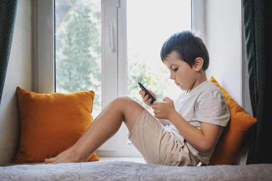 A 6-7 year old child sits on a windowsill at home with a mobile phone and plays. Mental health issues, unlimited gadgets, FOMO, harm to the child's psyche.