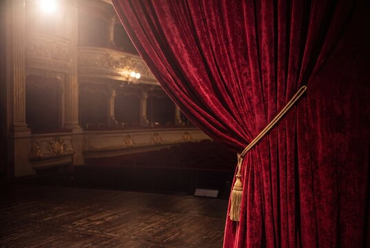 Luxurious red velvet theater curtain with golden tassel, tied back to reveal a dramatic spotlight on an empty, ornate stage within a classic opera house.