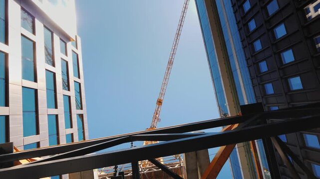 Low angle dynamic motion view of a massive construction crane between two modern buildings in an urban setting against clear blue sky.
