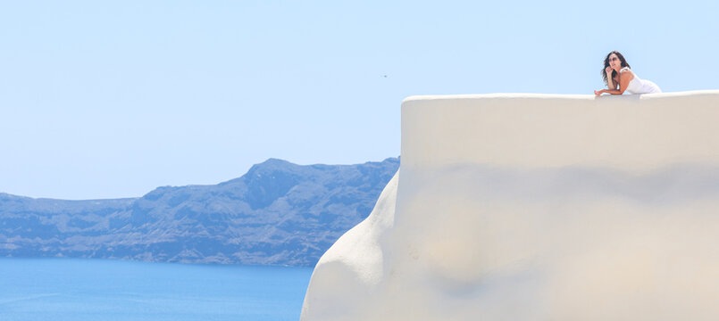 A woman in white rests on a sunlit terrace overlooking the Aegean Sea in Santorini, Greece, surrounded by bright Mediterranean light and serene coastal views.