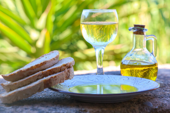 Bread, olive oil, and white wine sit on a stone table surrounded by greenery in Greece, evoking the warmth and simplicity of Mediterranean dining.