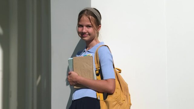 Teenage schoolgirl with backpack and books standing near building in profile and turning. Concept of education and back to school
