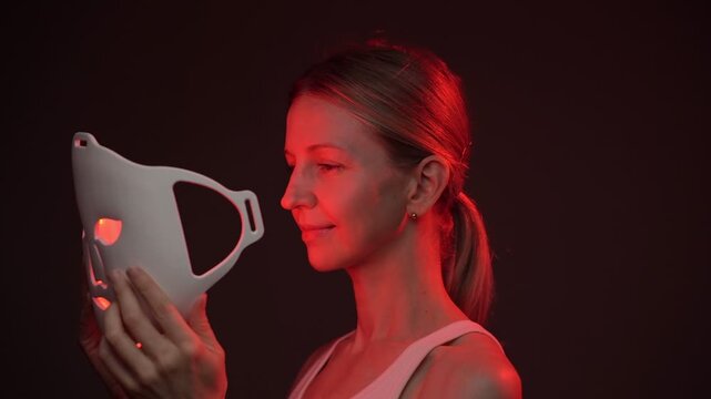 woman using an infrared facial mask. smiling and looking at camera.  Professional cosmetic skin care. vertical video footage black background studio shoot, contrast red light