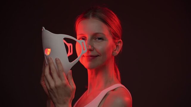 woman using an infrared facial mask. holding it face level. smiling and looking at camera.  Professional cosmetic skin care. vertical video footage black background studio shoot, contrast red light