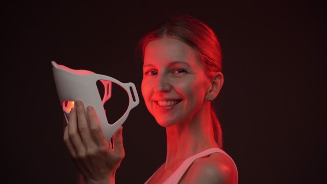 smiling woman holding infrared LED mask. biohacker theme red light professional skin care video footage. black background