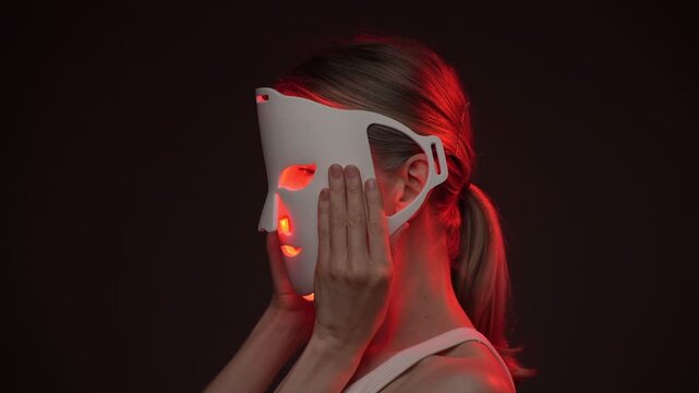 woman holding infrared LED mask puts it to her face, biohacker theme professional skin care video footage. black background