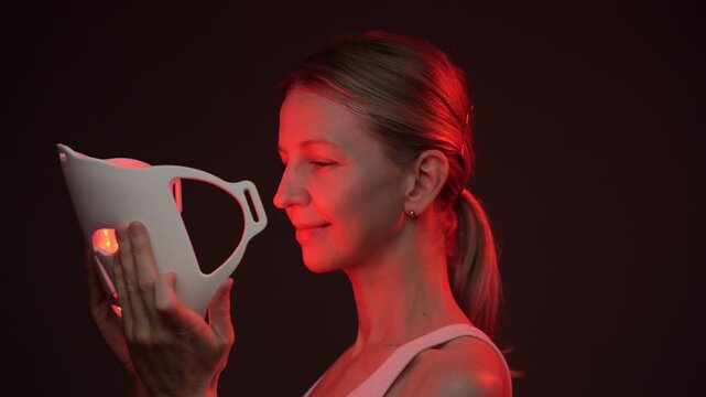 woman smiles while using an infrared facial mask. Professional cosmetic skin care. vertical video footage black background studio shoot, contrast red light
